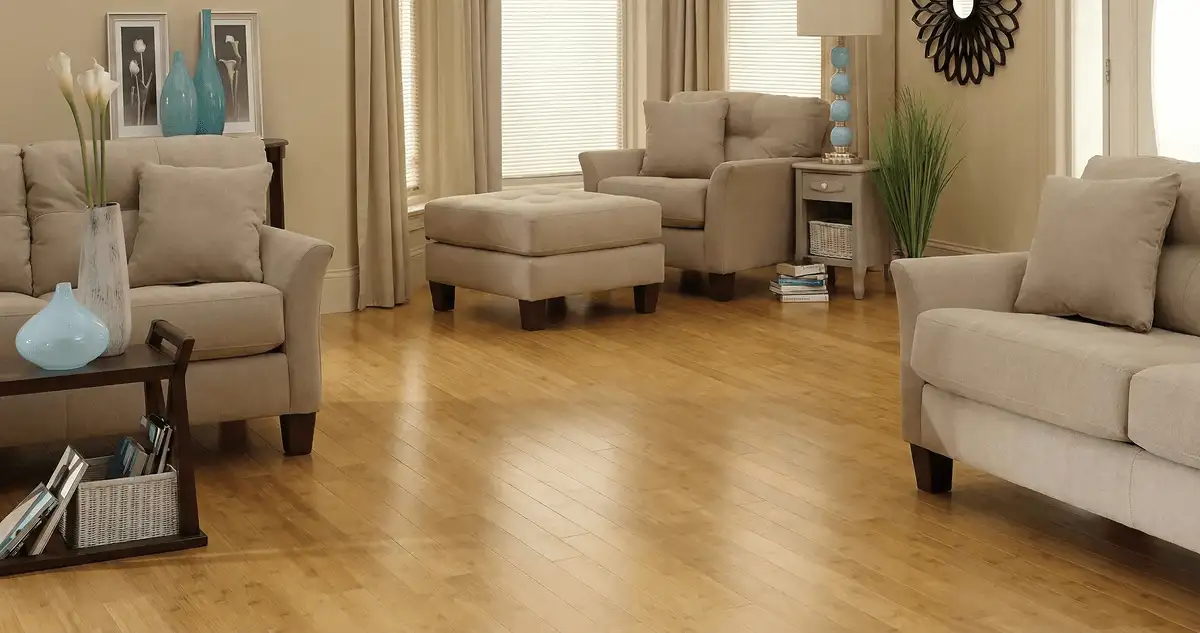 A living room with professionally installed bamboo flooring, beige sofas, a matching ottoman, a side table with a blue lamp, vases, framed pictures, and large windows with blinds and curtains in Providence, RI